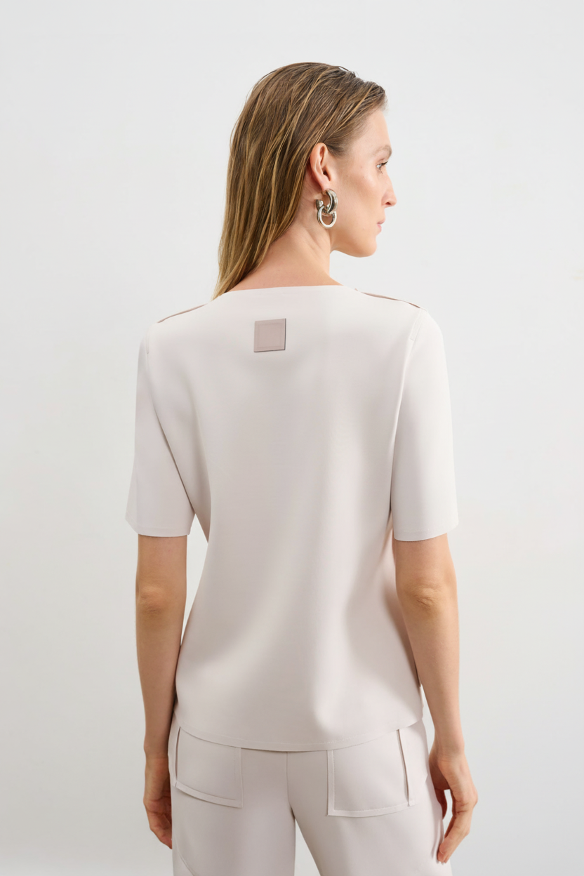 Modelo de costas usando uma blusa branca de mangas curtas com uma etiqueta retangular decorativa centralizada na parte superior, combinada com uma calça da mesma cor.