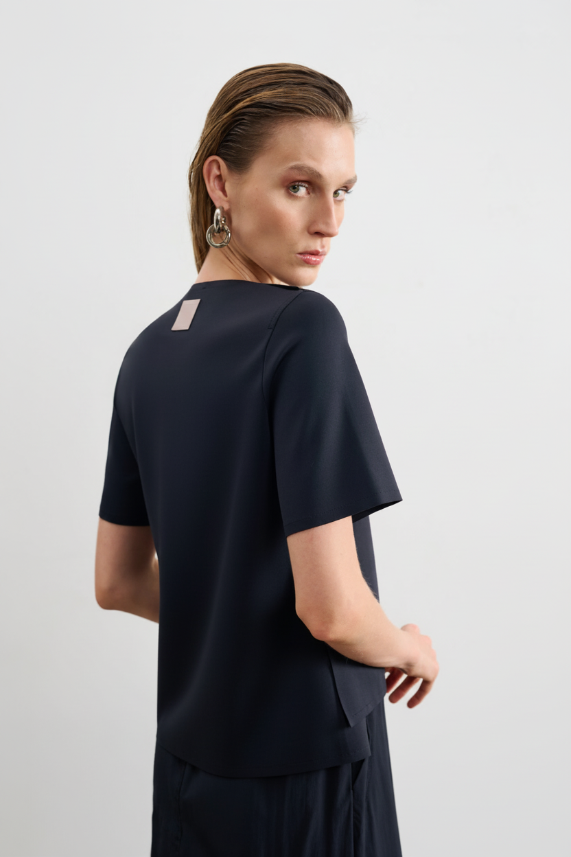 Modelo de costas usando uma blusa preta de mangas curtas com uma etiqueta retangular decorativa centralizada na parte superior, combinada com uma saia da mesma cor.