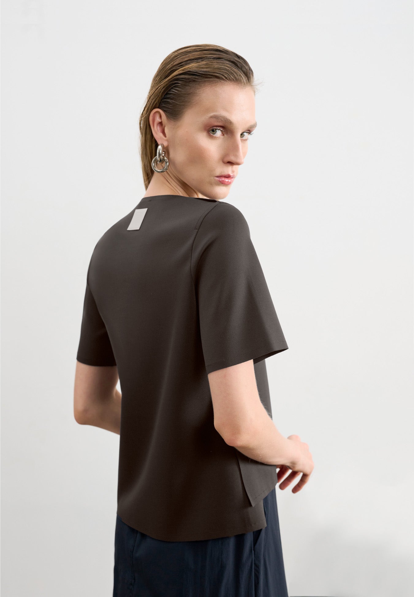 Modelo de costas usando uma blusa marrom de mangas curtas com uma etiqueta retangular decorativa centralizada na parte superior, combinada com uma saia preta.