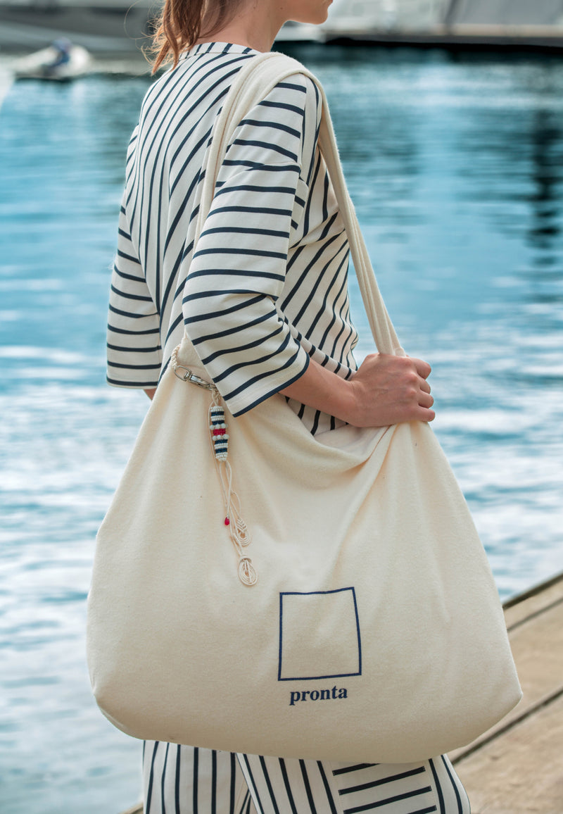 Modelo de costas carrega uma bolsa off-white com logomarca e um chaveiro artesanal pendurado, vestindo um conjunto listrado.