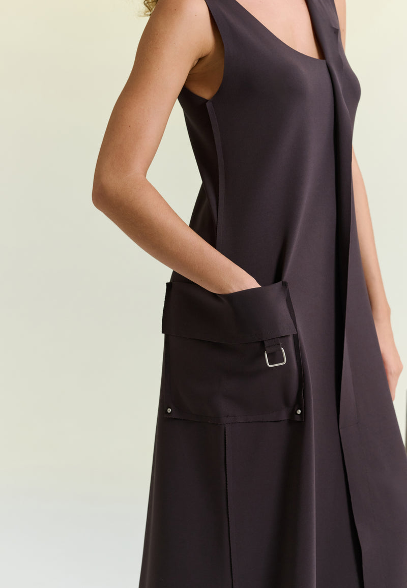 Modelo com vestido midi marrom sem mangas com recorte frontal, bolso lateral com argola de metal.