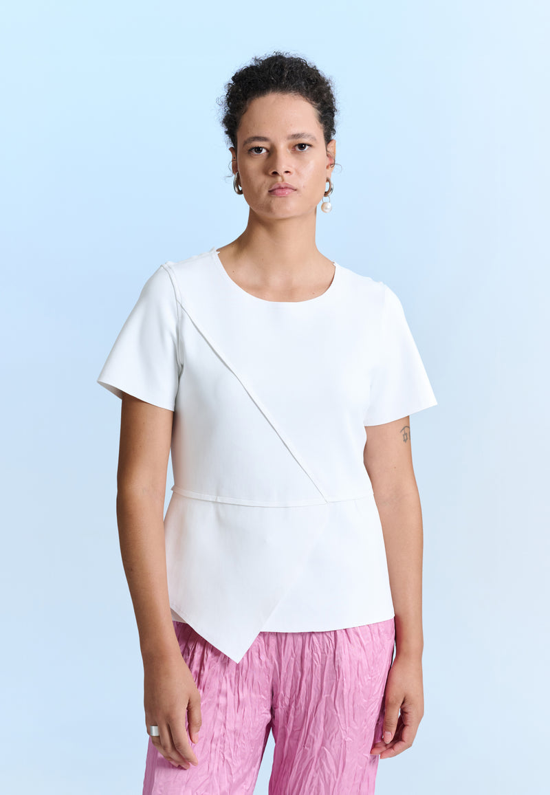Modelo veste uma blusa branca de mangas curtas com transpasse frontal, combinada com uma calça rosa de textura plissada.