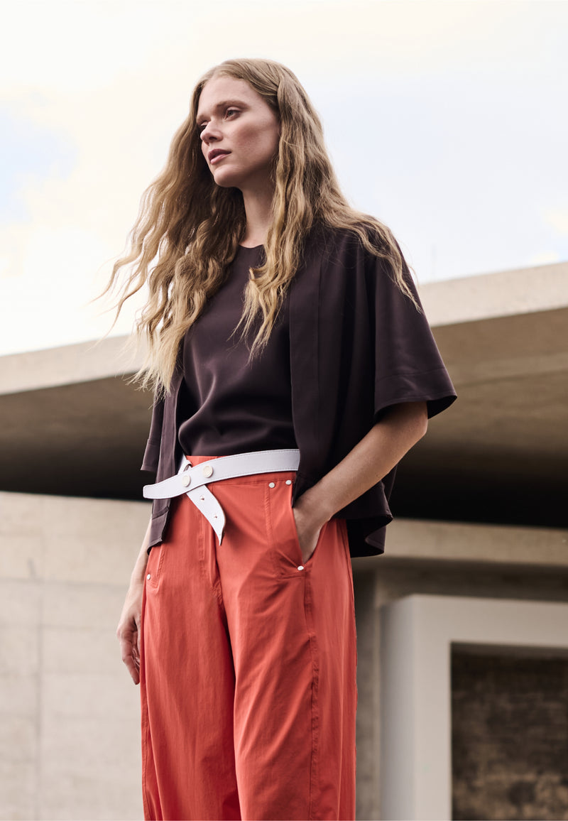 Modelo veste sobreposição marrom de mangas 3/4 e blusa marrom, combinando com calça de alfaiataria terracota com rebites e cinto branco.