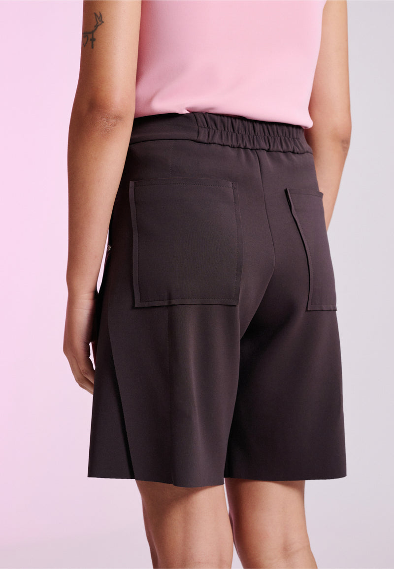 Modelo de costas exibe uma bermuda marrom com cós elástico e bolsos quadrados aplicados.