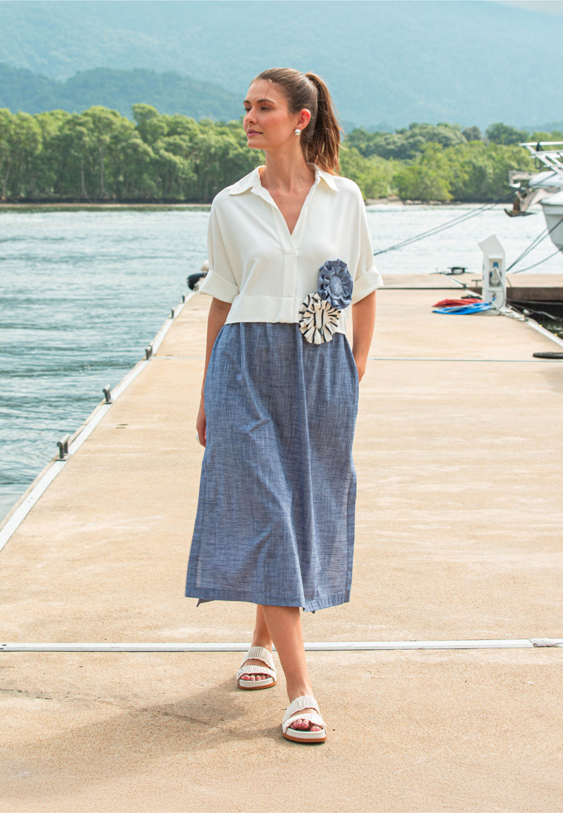 Modelo usando um vestido de comprimento midi que combina uma parte superior estilo camisa branca com flor decorativa no busto e uma saia marinho mescla de caimento fluido.