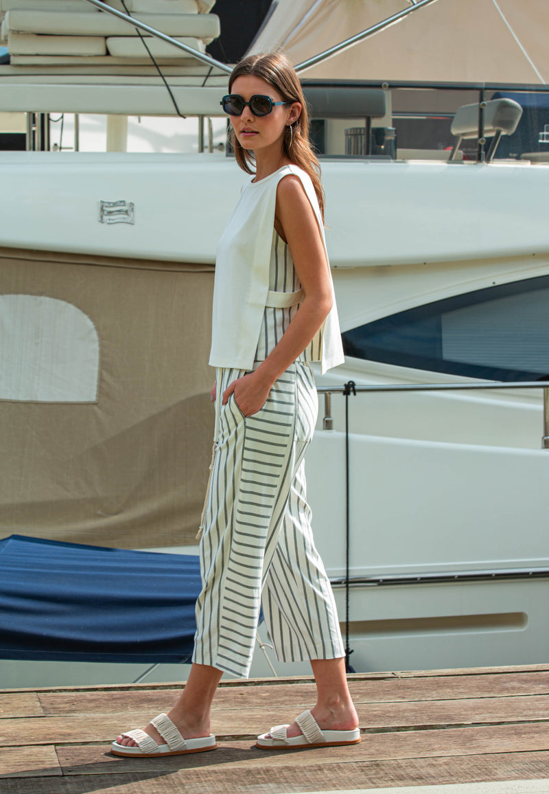 Modelo de perfil com conjunto casual composto por uma blusa branca com recorte lateral com listras verticais e calça listrada em cinza.