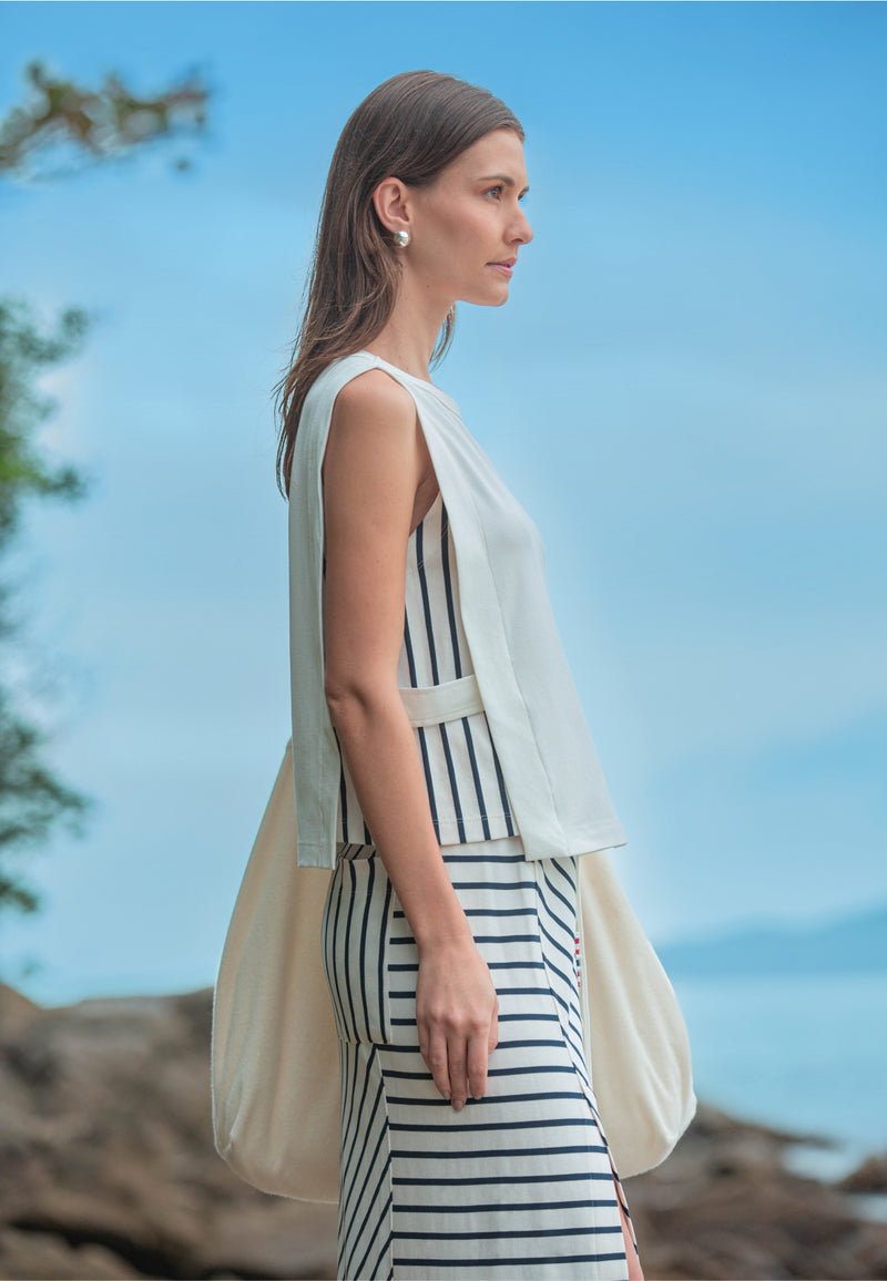 Modelo de perfil usando um conjunto de blusa branca com laterais listrada e saia listrada, acompanhado de uma bolsa lateral volumosa.