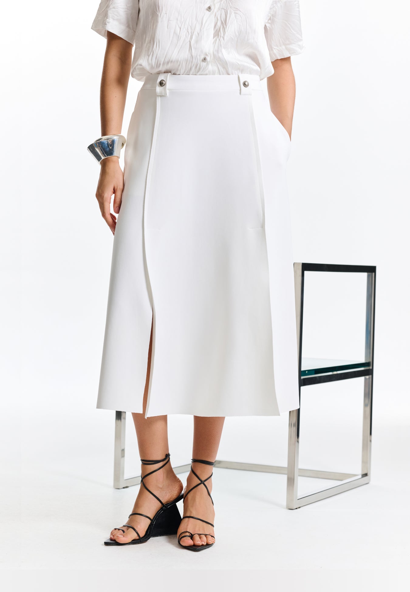 Modelo veste saia midi branca de modelagem evasê, destacando os passantes com botões metálicos decorativos e a combinação com sandálias pretas de tiras e salto geométrico.