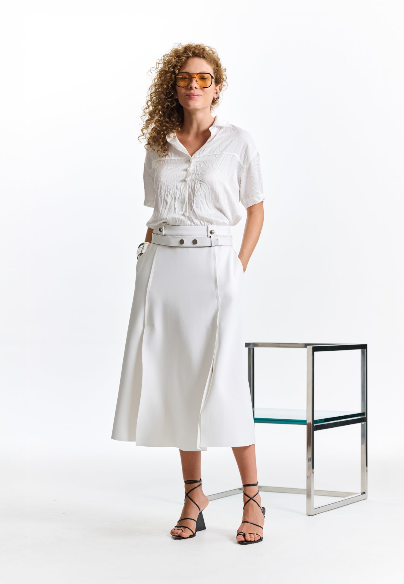Modelo veste uma camisa branca texturizada com botões e uma saia midi branca com recortes, cinto branco e sandálias pretas de tiras.