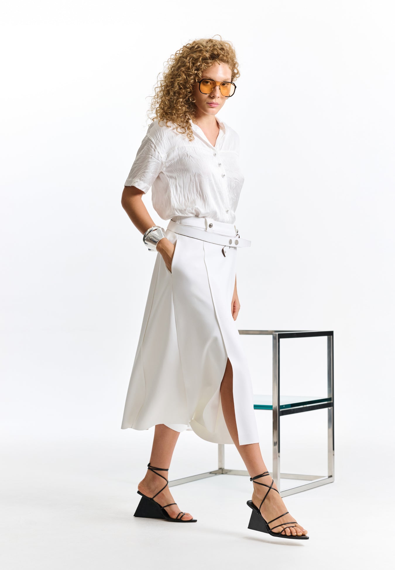 Modelo veste uma camisa branca texturizada com botões e uma saia midi branca com recortes, cinto branco e sandálias pretas de tiras.
