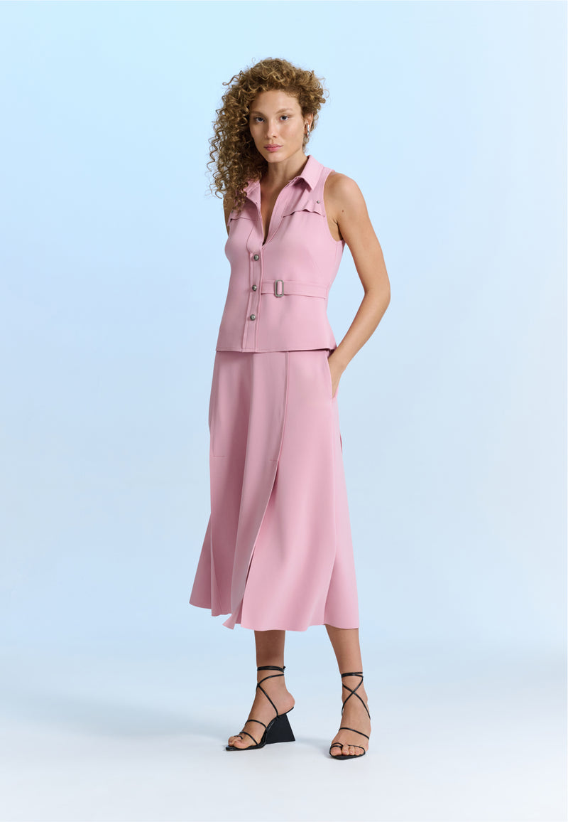 Modelo veste um conjunto rosa composto por um colete com abotoamento frontal e detalhes de fivelas metálicas, combinado com uma saia midi evasê e sandálias pretas de tiras.