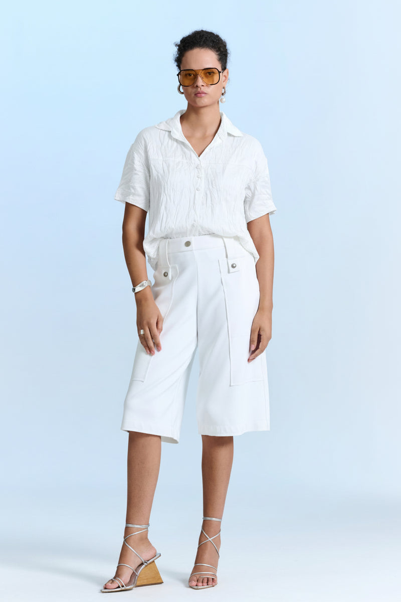 Modelo veste uma camisa branca texturizada de mangas curtas e bermuda de alfaiataria branca com botões metálicos, calçando sandálias pratas de salto geométrico.