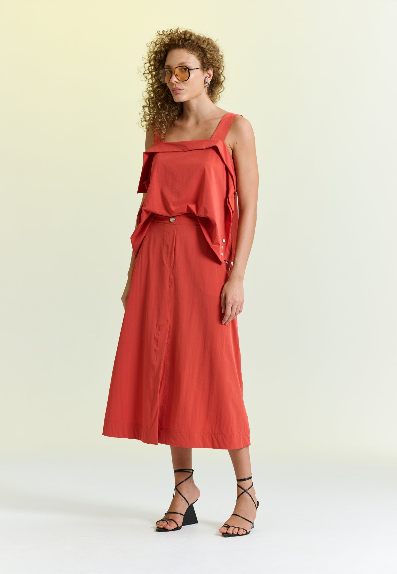 Modelo veste uma blusa terracota com alças largas e detalhe de sobreposição estruturada no decote, combinada com saia midi da mesma cor e sandálias pretas de salto geométrico.