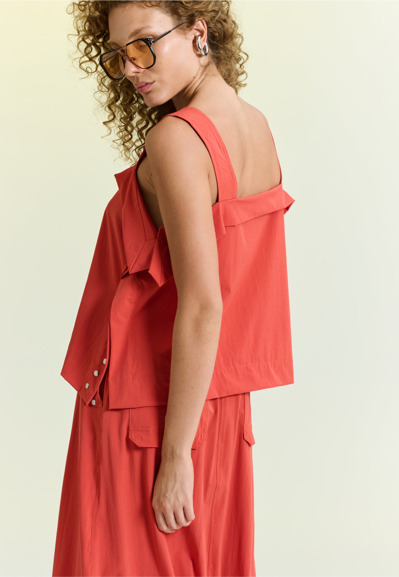 Modelo de costas exibe blusa com alças largas e sobreposição no decote.