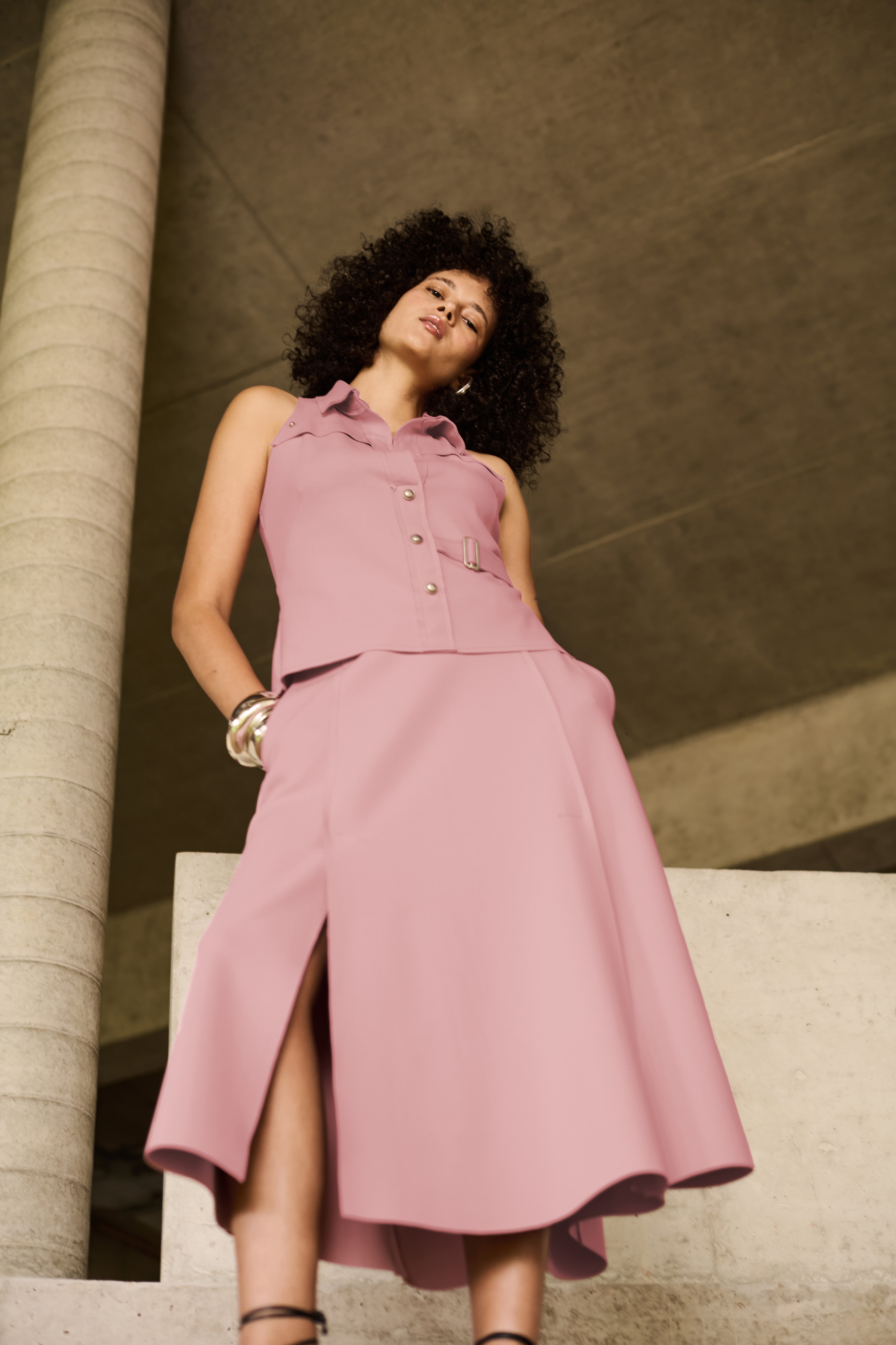 Modelo veste colete rosa com gola colarinho, abotoamento frontal por botões metálicos, e detalhe de cinto com fivela na cintura, combinando com saia midi rosa.