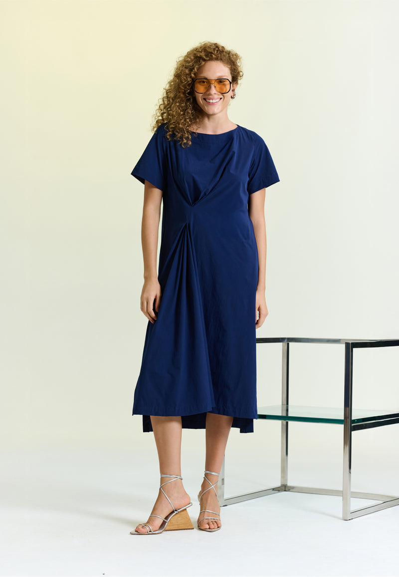 Modelo usa um vestido midi azul de mangas curtas e decote canoa, apresentando um detalhe de prega lateral e sandálias de salto geométrico. 