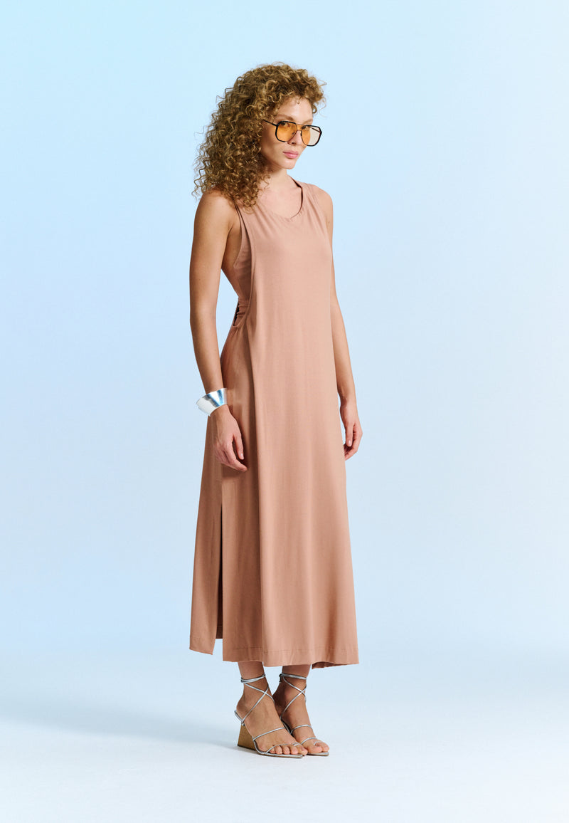 Modelo usa um vestido midi bege sem mangas com decote arredondado e fendas laterais, acompanhado sandálias de tiras prateadas.