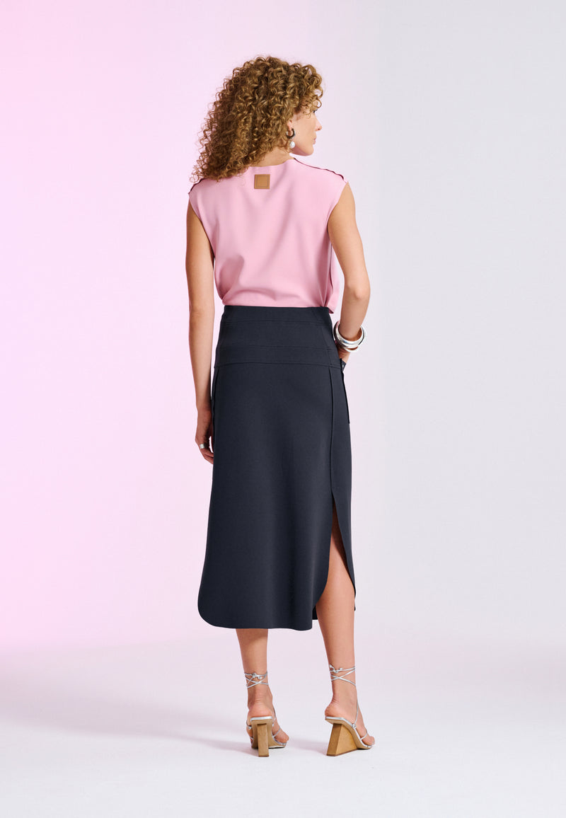 Modelo de costas exibe uma blusa rosa com etiqueta na gola e uma saia midi preta de cintura alta com fenda na lateral, calçando sandálias pratas de salto geométrico.