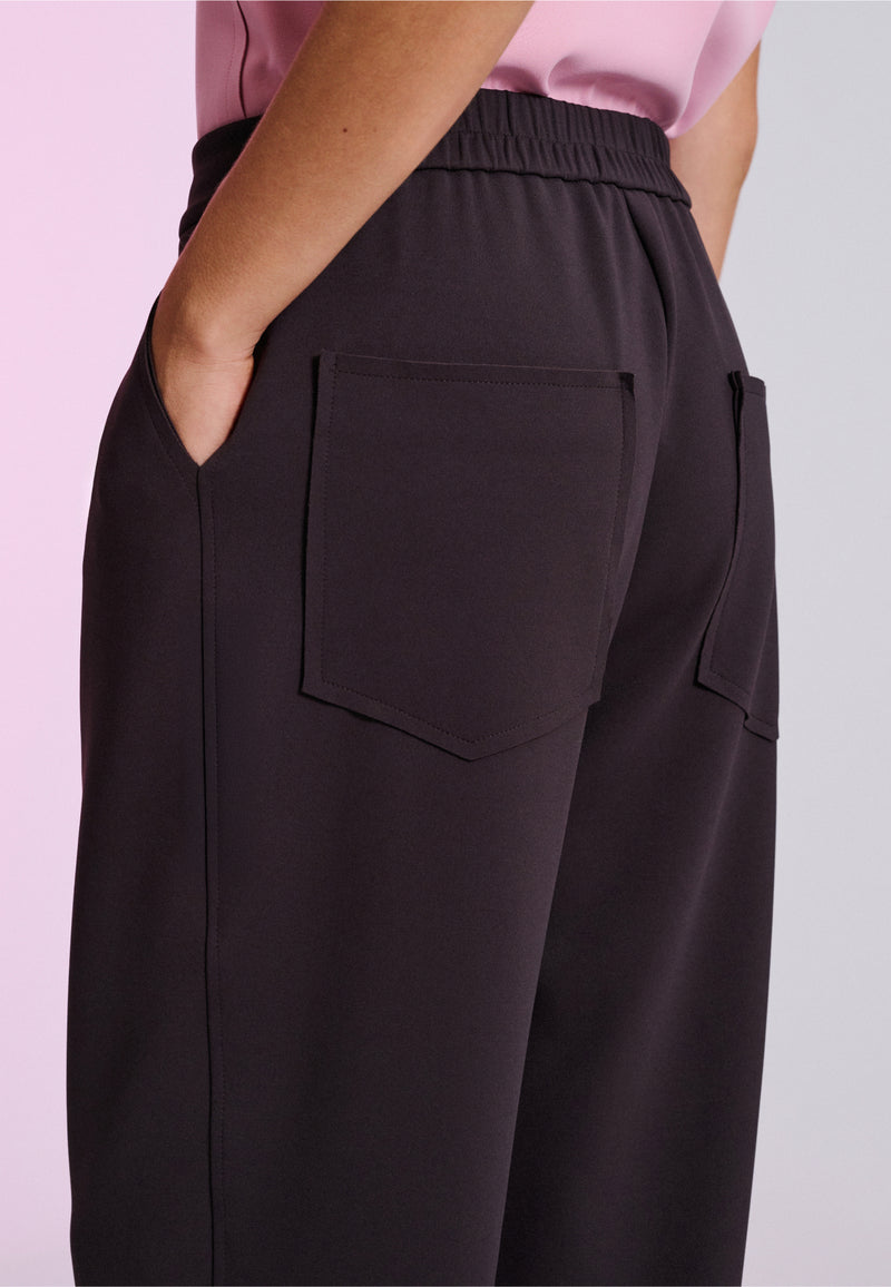 Modelo de costas exibe calça de alfaiataria marrom com cós elástico, bolsos traseiros quadrados sobrepostos.
