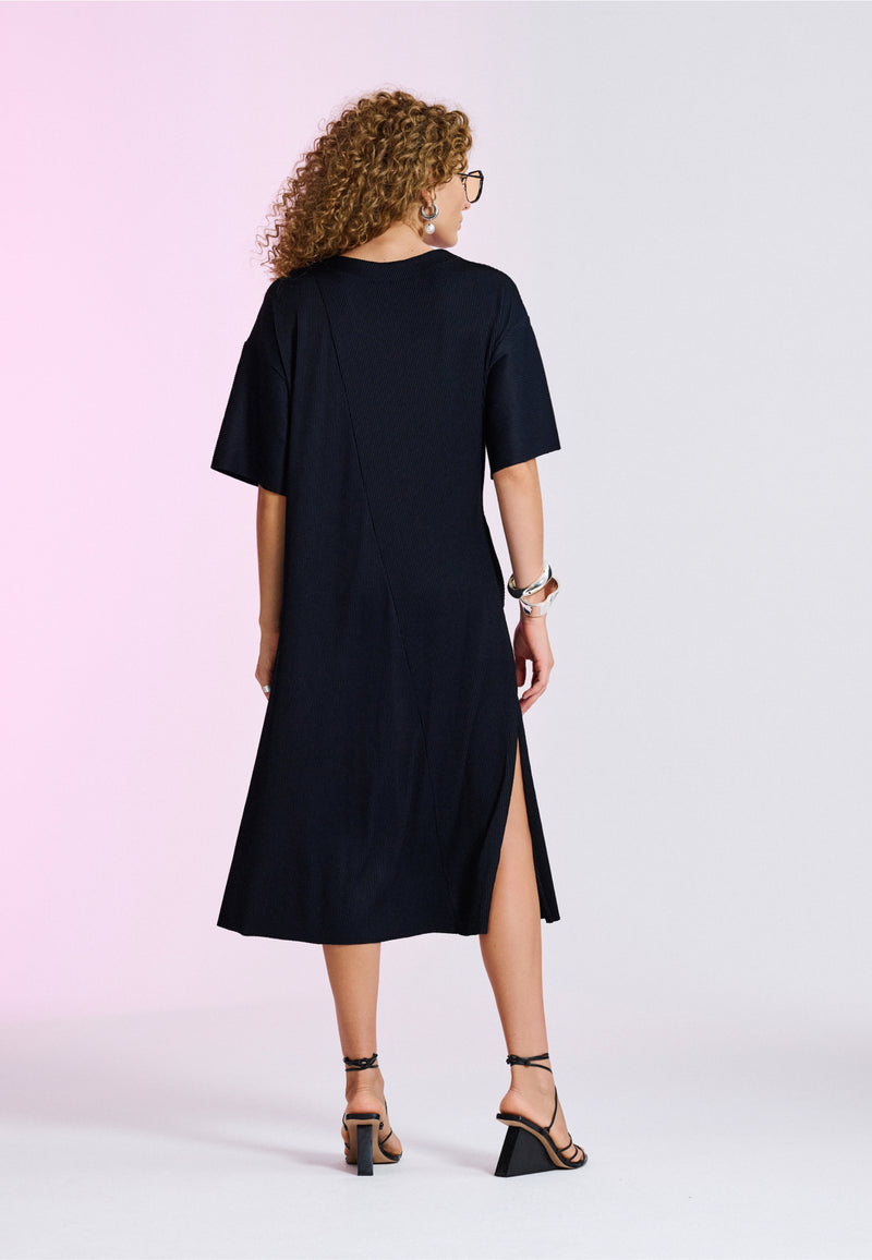 Modelo de costas exibe um vestido midi preto de textura plissada com fenda lateral, acompanhado por uma sandálias de tiras com salto geométrico.