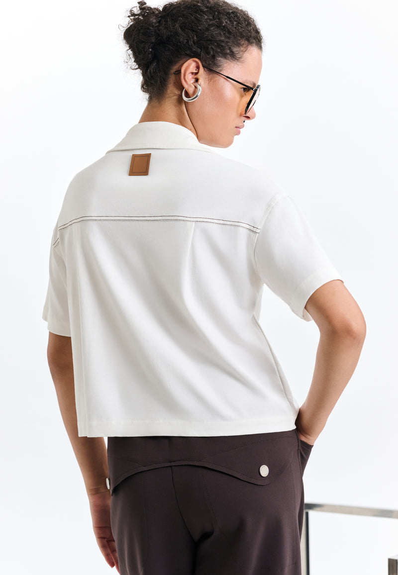 Modelo de costas exibe camisa branca de mangas curtas com etiqueta na pala, combinando com calça marrom que apresenta lapelas pontiagudas e botões de metal.
