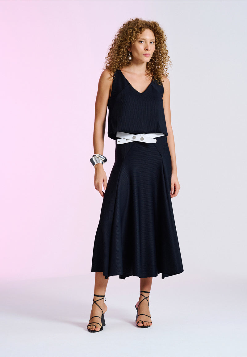 Modelo veste um conjunto preto texturizado de blusa com decote em V e saia midi, destacando-se por um cinto branco largo e sandálias pretas de tiras.