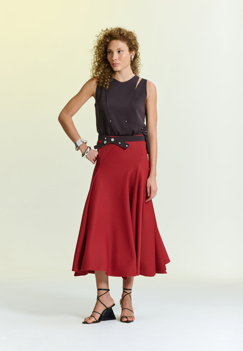 Modelo veste uma blusa marrom sem mangas com detalhes vazados no ombro e uma saia midi vermelha texturizada,destacando-se por um cinto marrom largo e sandálias pretas de tiras.