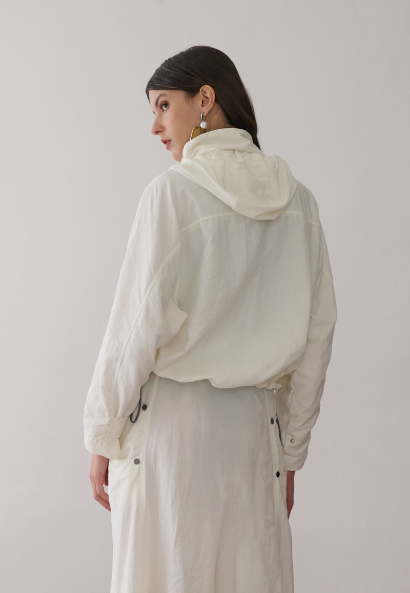 Modelo de costas veste uma jaqueta off-white com capuz, combinada com uma saia midi off-white que possui bolsos laterais utilitários com rebites.