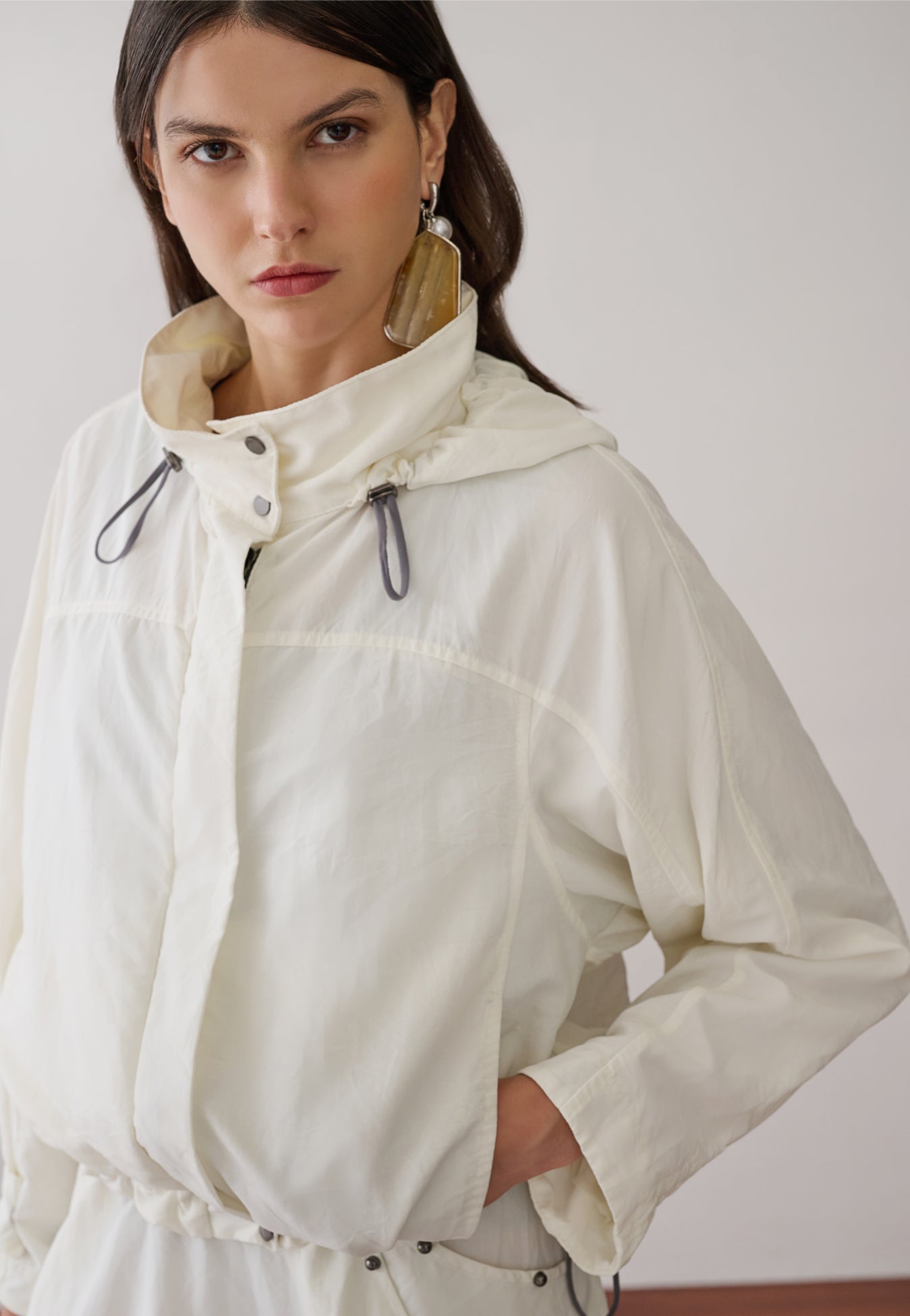 Modelo veste uma jaqueta off-white com capuz e cordões de ajuste, apresentando detalhes de botões de pressão e recortes pespontados.