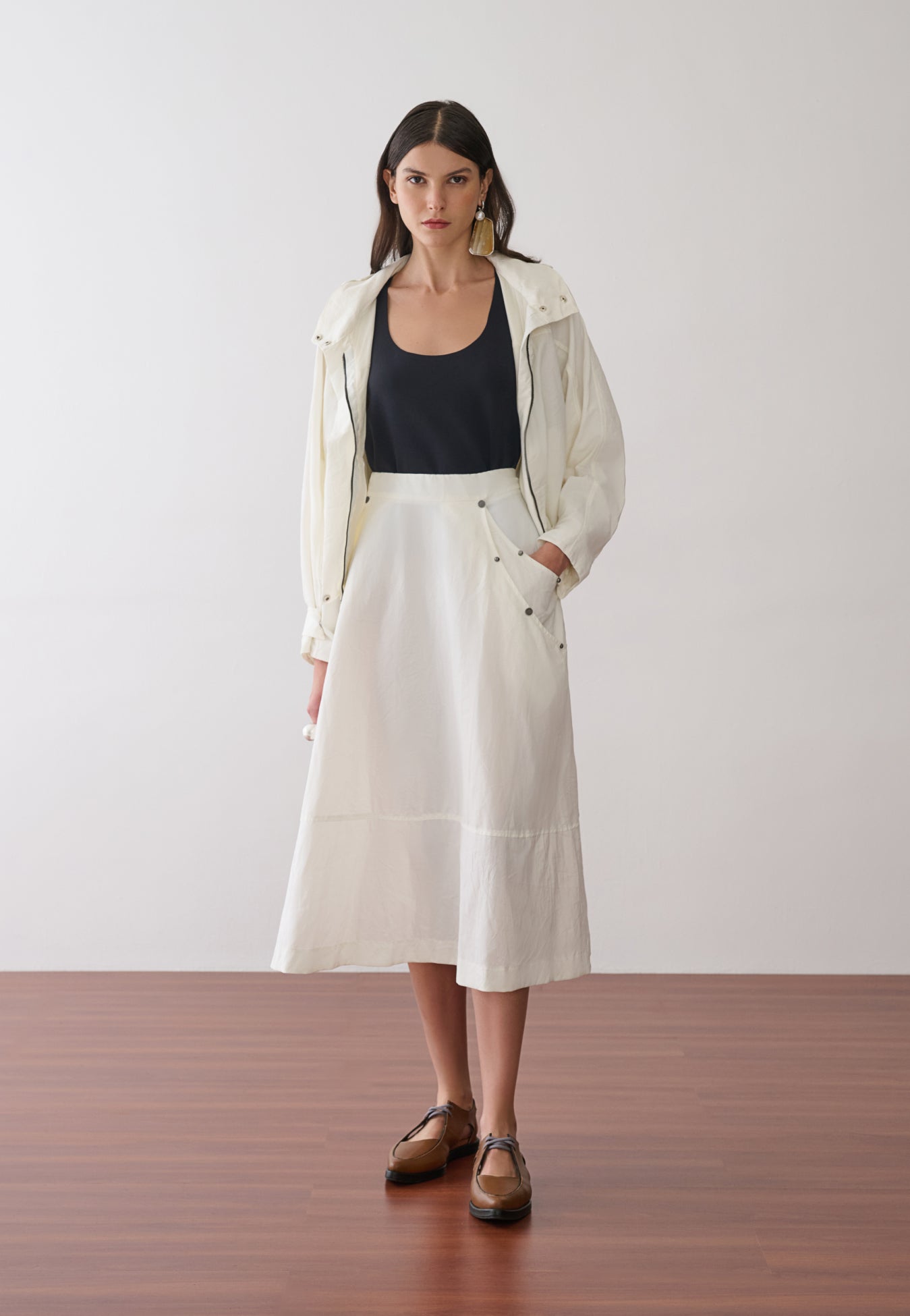 Modelo veste uma jaqueta off-white com capuz sobre uma regata preta, combinada com uma saia midi off-white que possui bolsos laterais utilitários com rebites e sapatos marrons de bico fino.