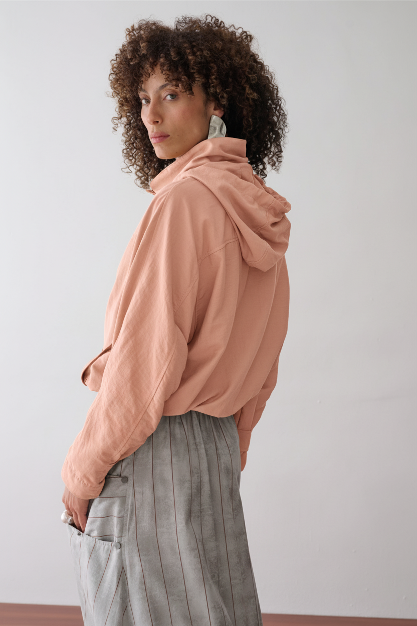 Modelo de costas veste uma jaqueta na cor pêssego com capuz, combinada com uma saia midi cinza com listras marrons que possui bolsos laterais utilitários com rebites.