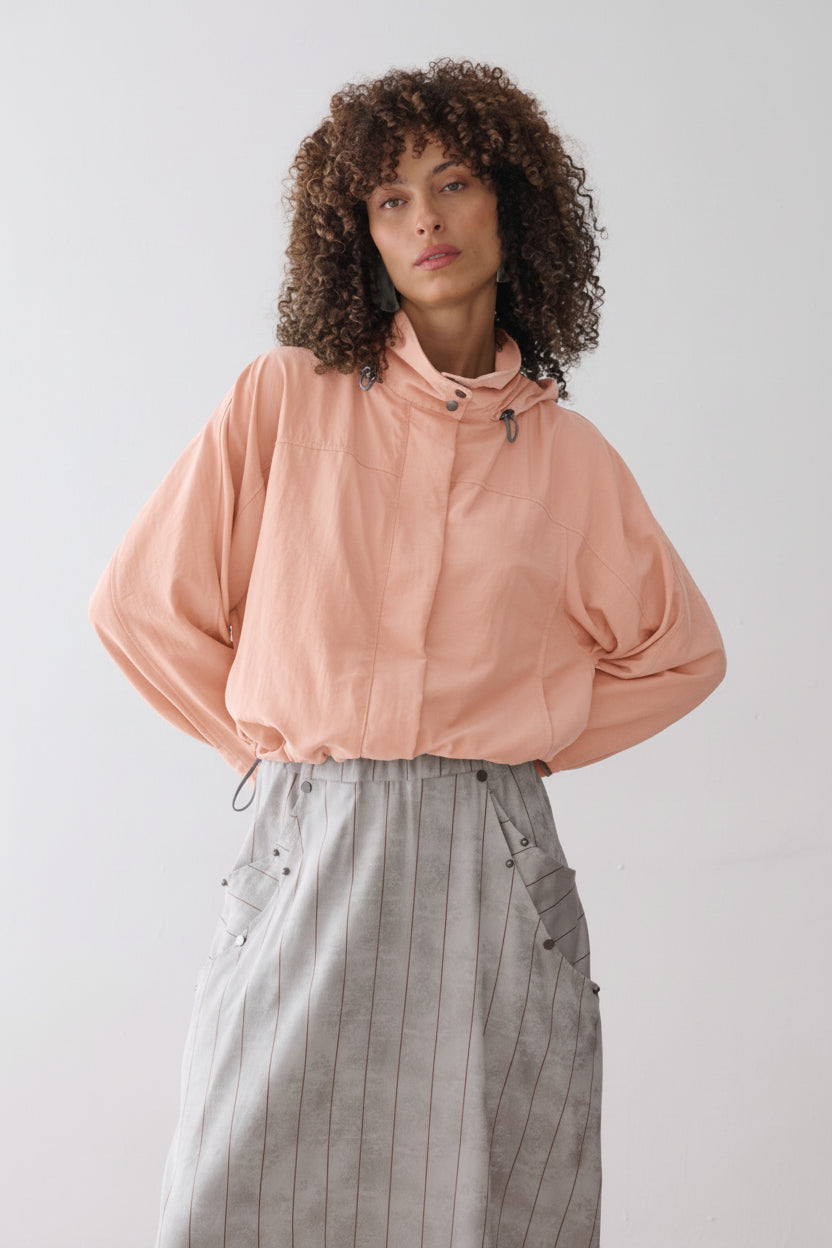 Modelo veste uma jaqueta na cor pêssego com capuz, combinada com uma saia midi cinza com listras marrons que possui bolsos laterais utilitários com rebites.