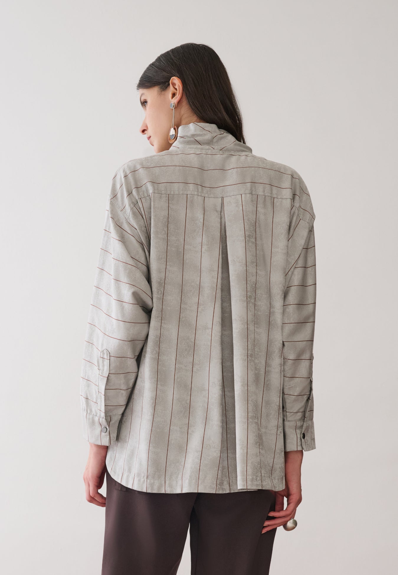 Modelo de costas veste uma camisa cinza com listras marrons e prega centralizada, combinada com calças marrons. 