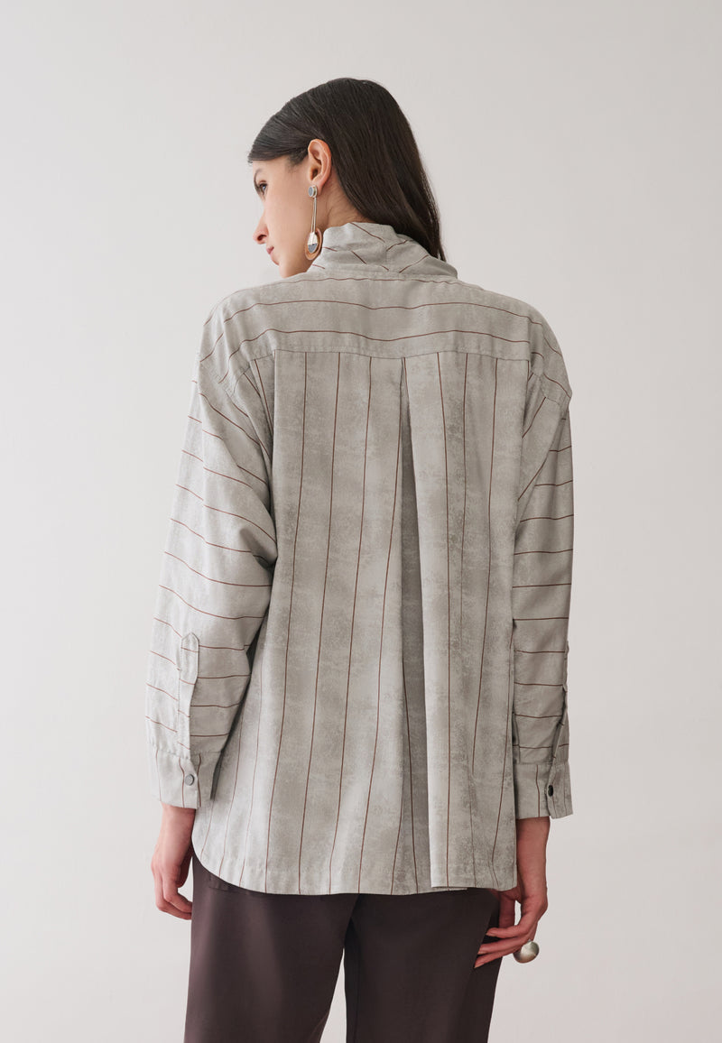 Modelo de costas veste uma camisa cinza com listras marrons e prega centralizada, combinada com calças marrons. 