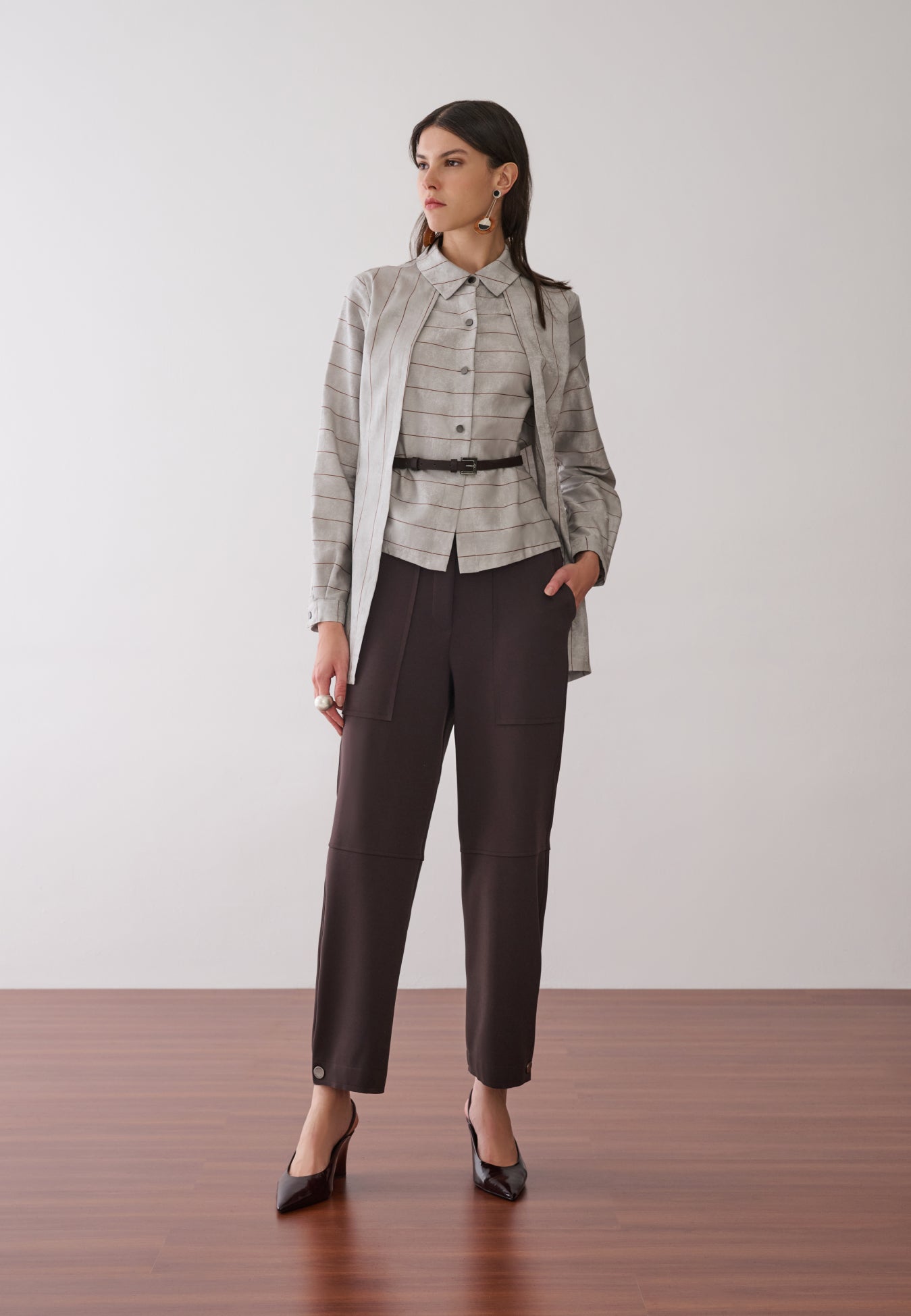 Modelo veste uma camisa com sobreposição integrada cinza com listras marrons, combinada com calças marrons de bolsos utilitários e cinto fino e sapatos escuros de bico fino.