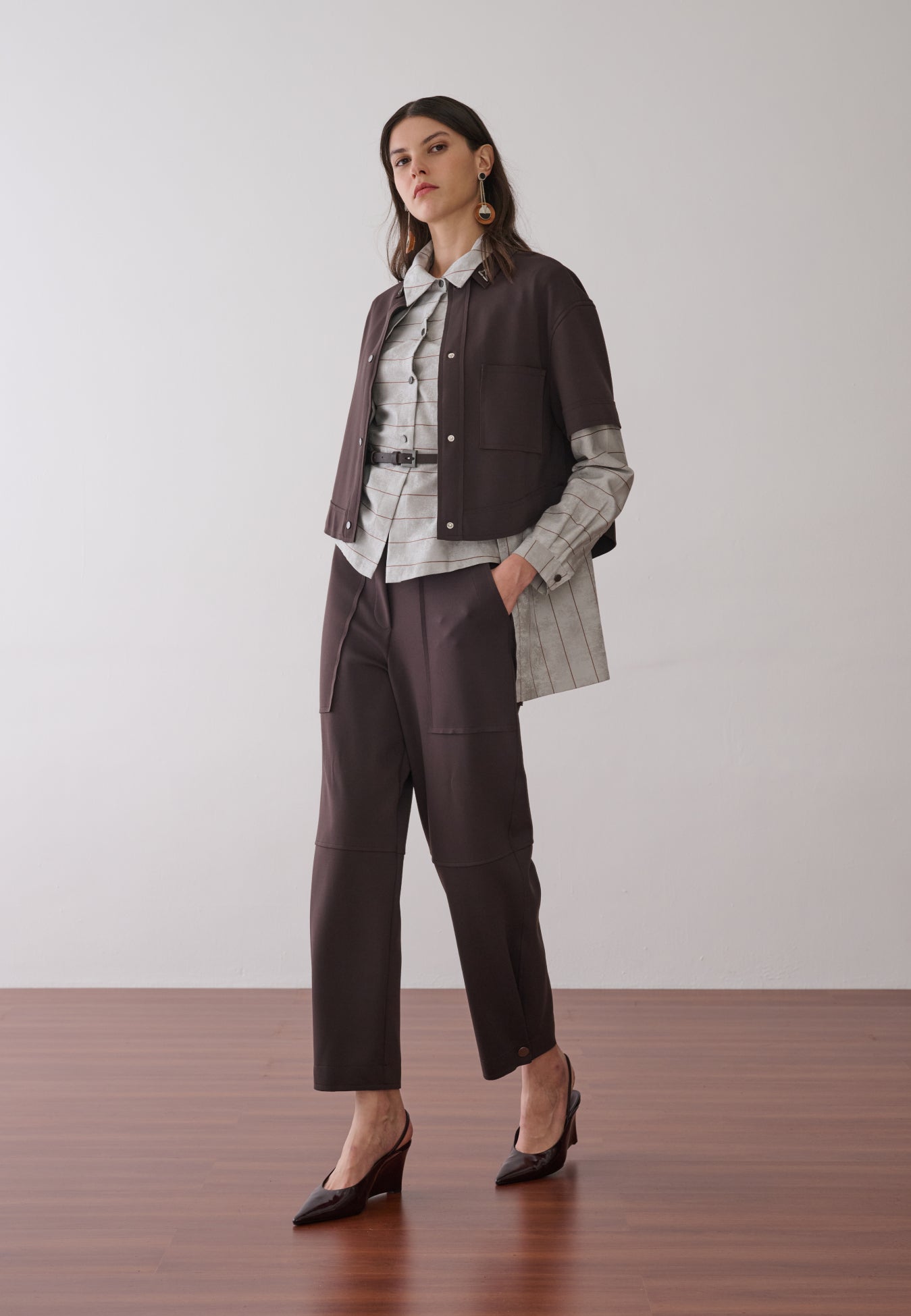 Modelo veste uma camisa com sobreposição integrada cinza com listras marrons, combinada com colete e calça marrom de bolsos utilitários e cinto fino e sapatos escuros de bico fino.