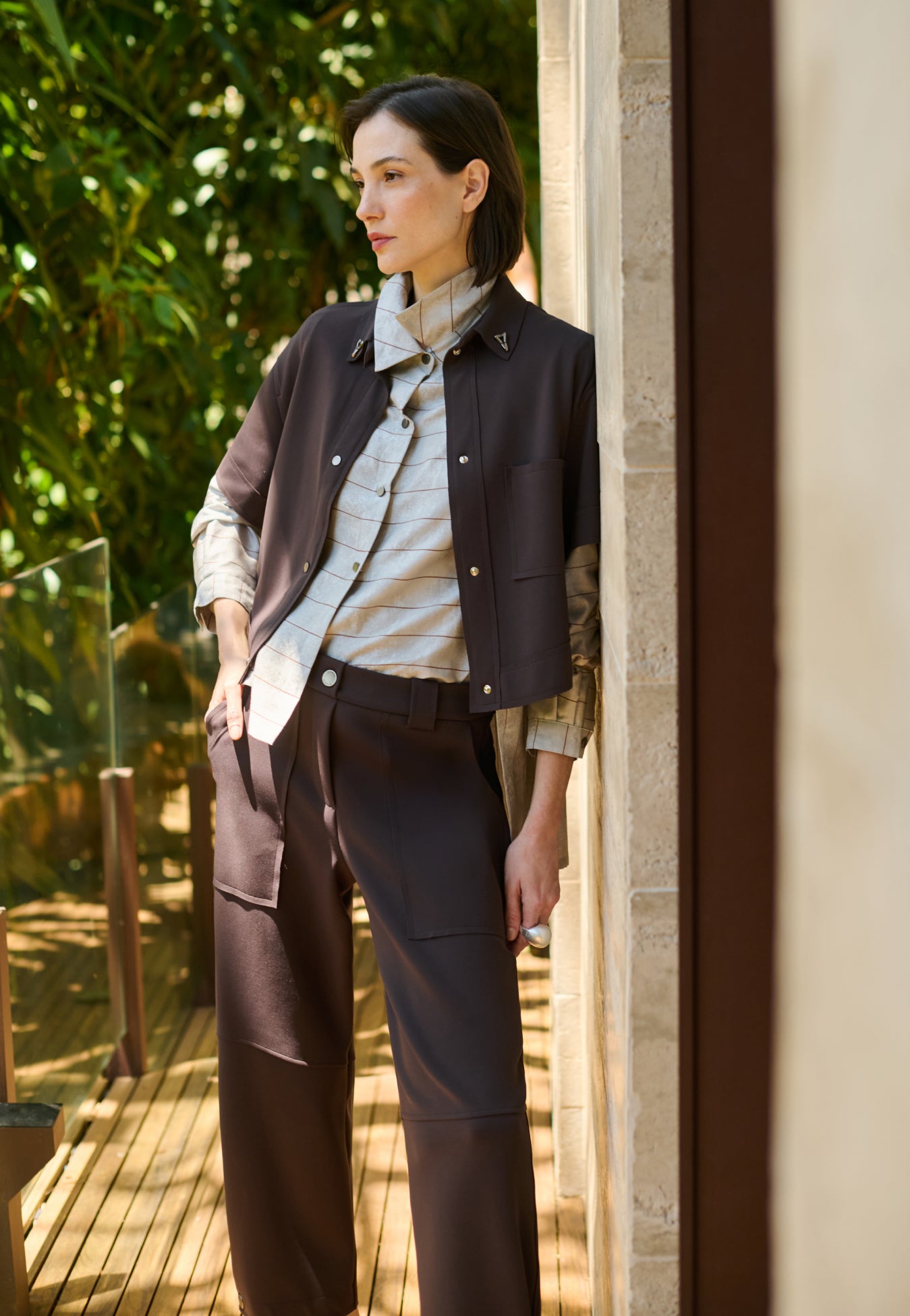 Modelo veste uma camisa com sobreposição integrada cinza com listras marrons, combinada com colete e calça marrom de bolsos utilitários.