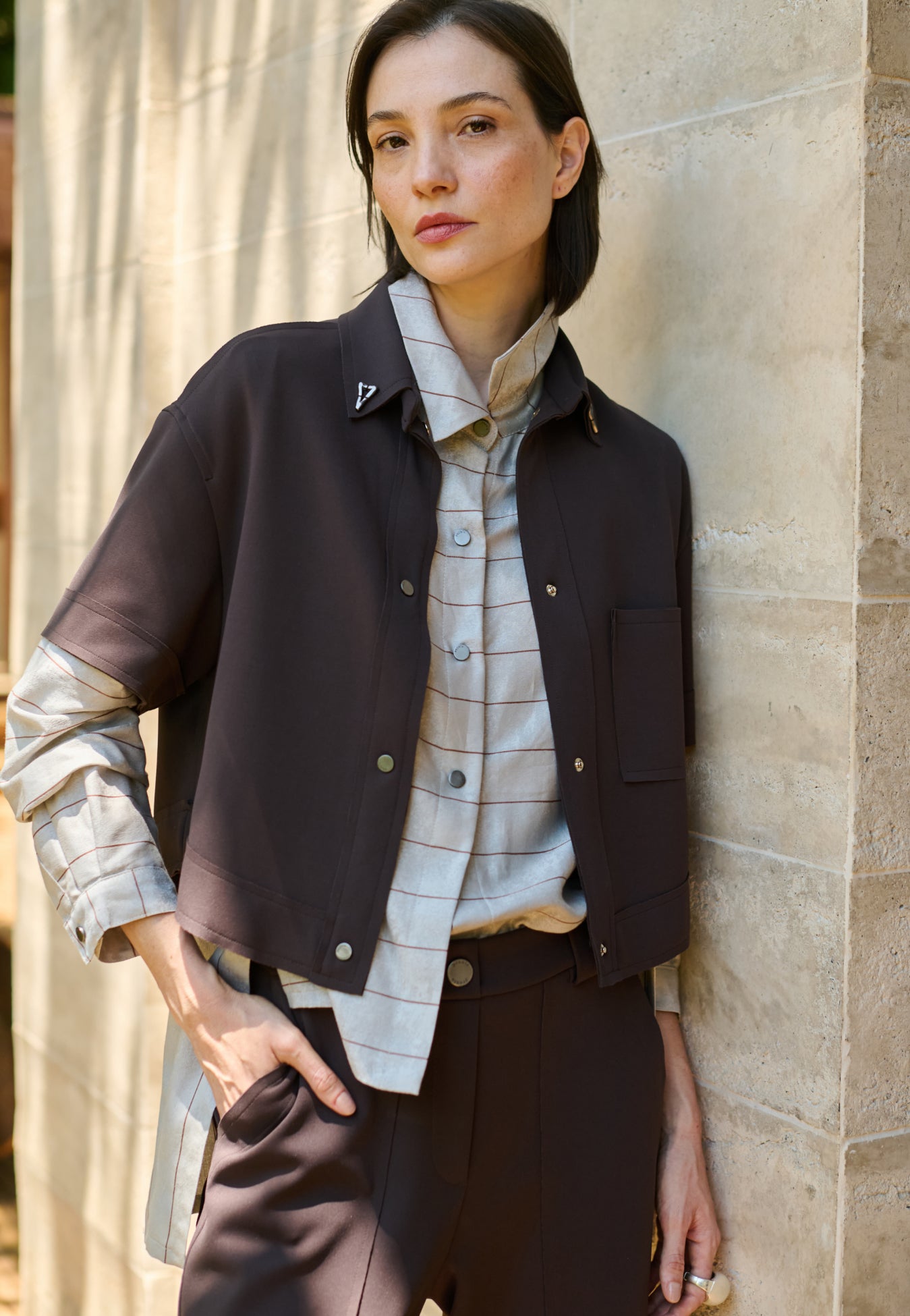 Modelo veste uma camisa com sobreposição integrada cinza com listras marrons, combinada com colete e calça marrom de bolsos utilitários.