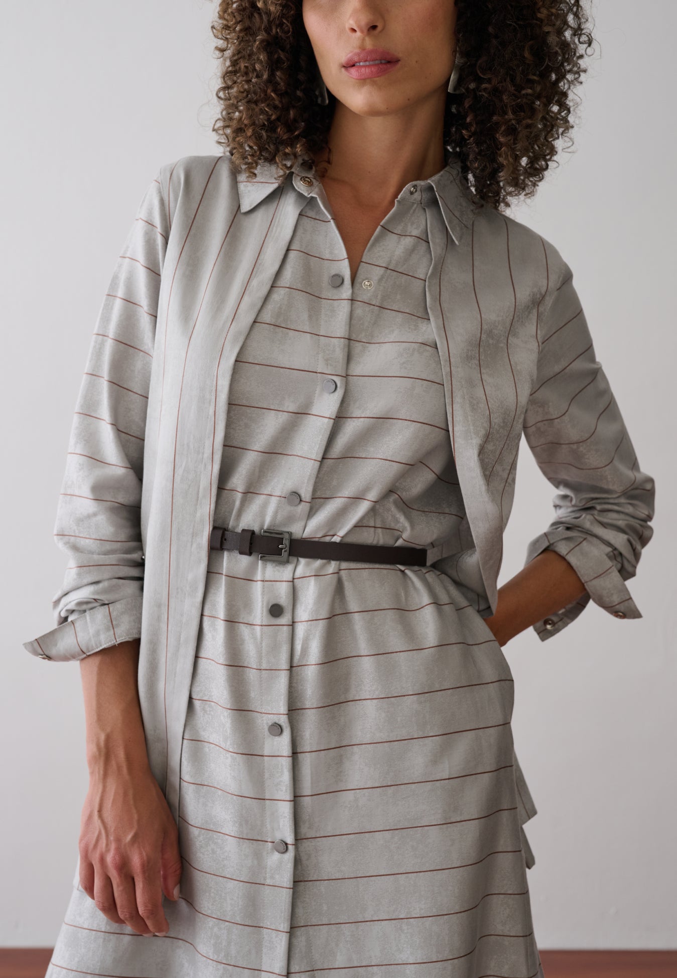Modelo usa um vestido midi com sobreposição integrada em cinza e listras marrons, abotoamento frontal e acinturado por um cinto fino preto.