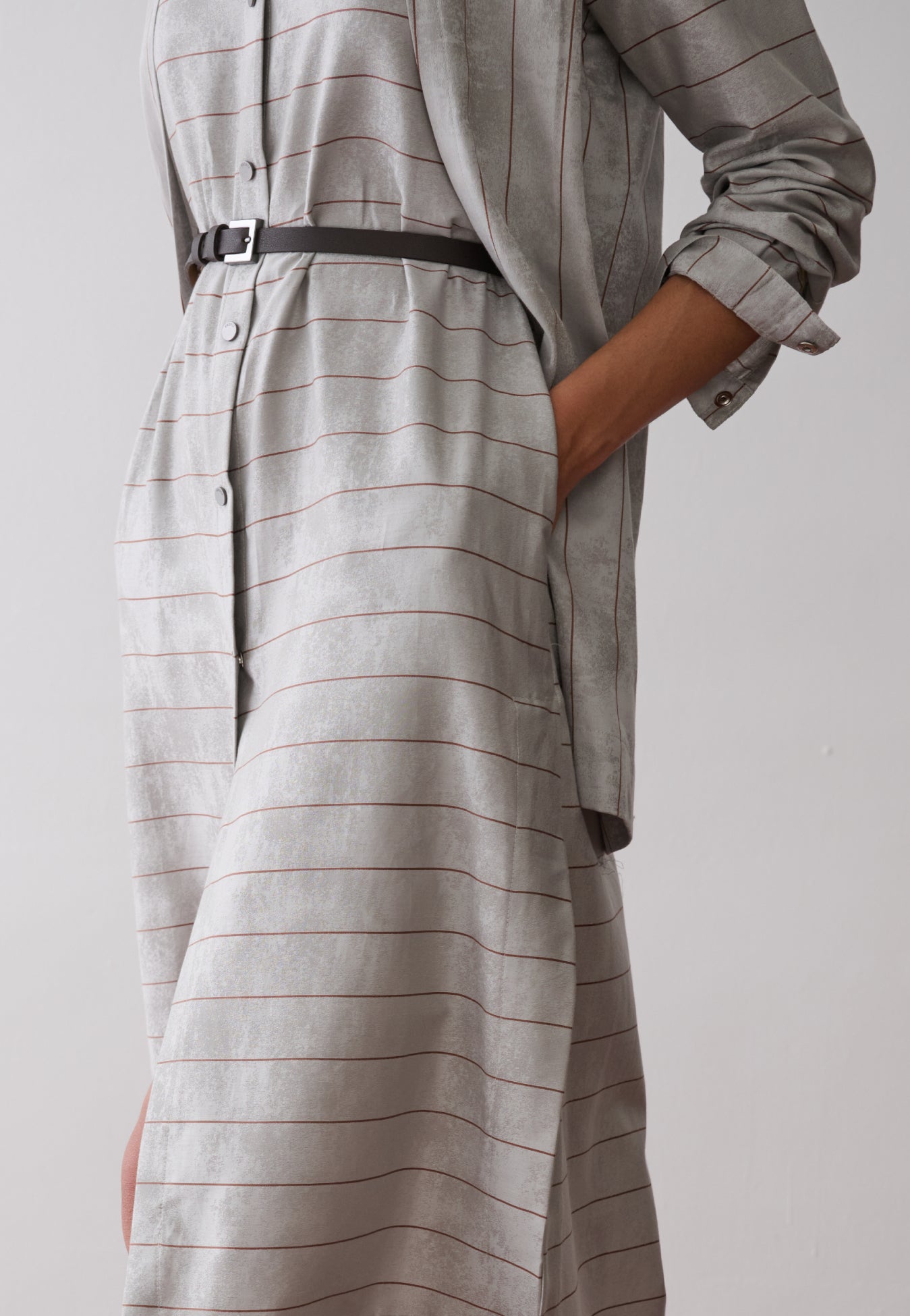 Modelo usa um vestido midi com sobreposição integrada em cinza e listras marrons, abotoamento frontal e acinturado por um cinto fino preto.