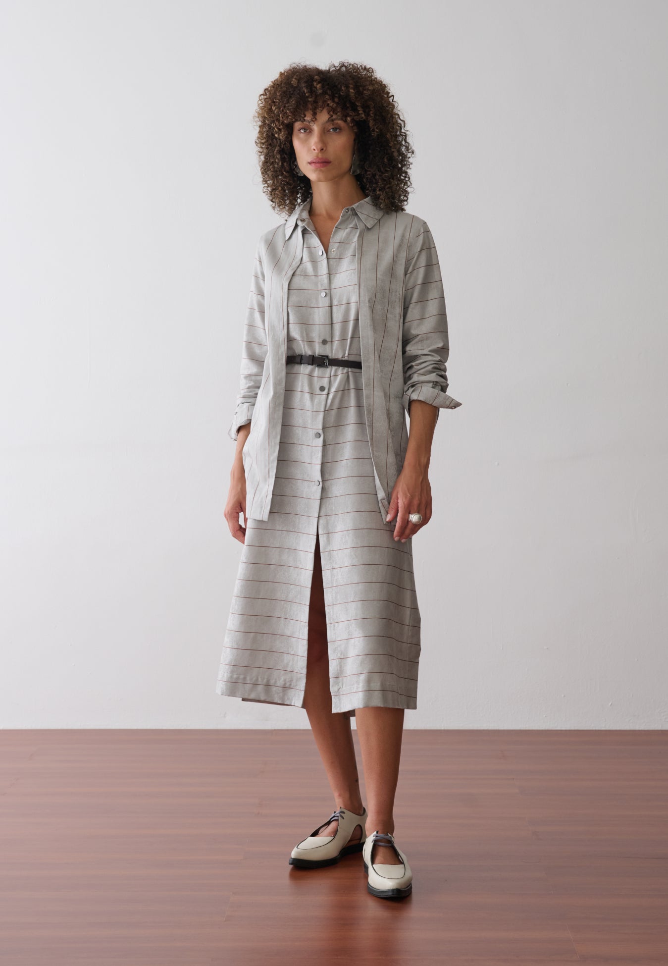 Modelo usa um vestido midi com sobreposição integrada em cinza e listras marrons, abotoamento frontal e acinturado por um cinto fino preto.