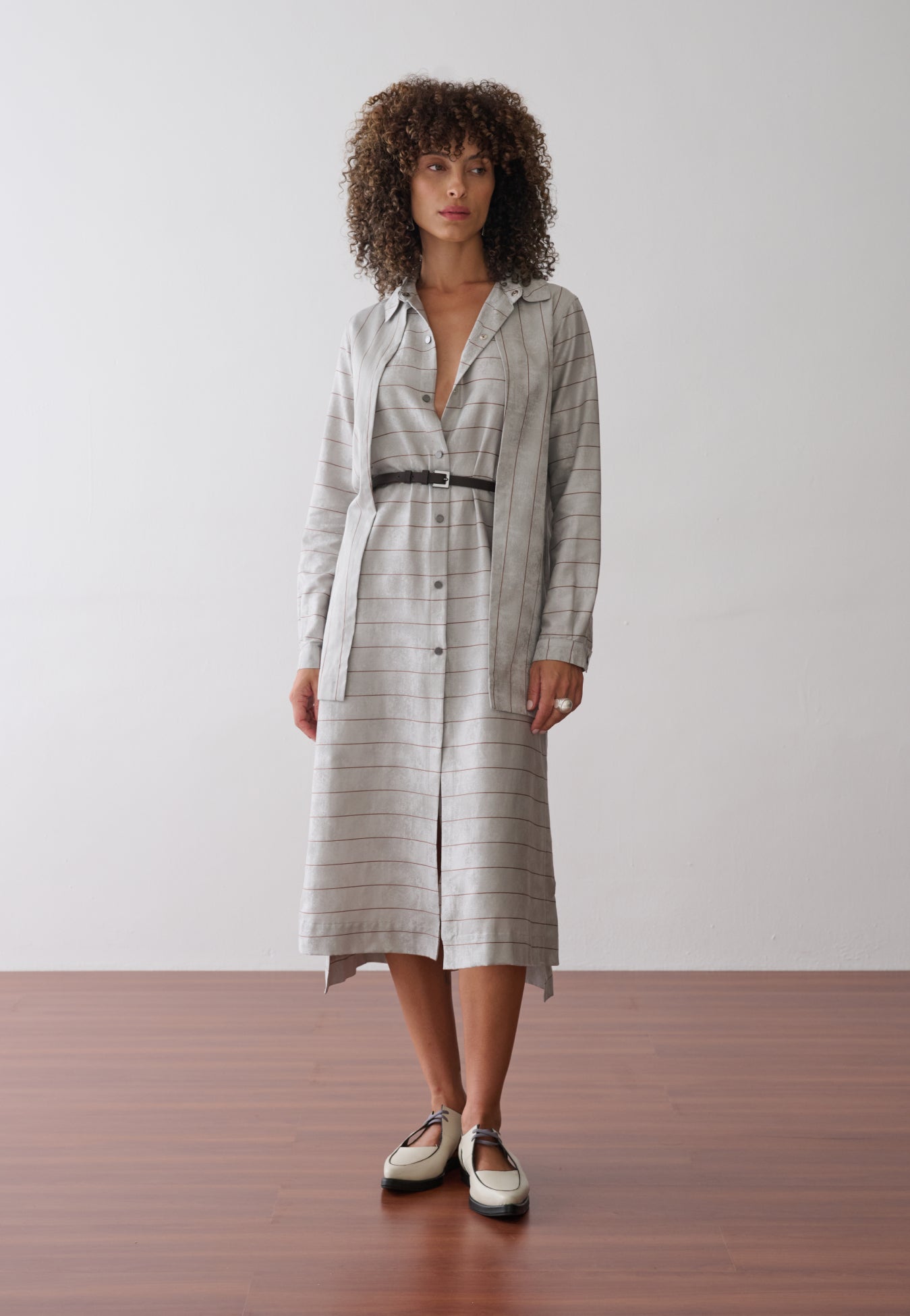 Modelo usa um vestido midi com sobreposição integrada em cinza e listras marrons, abotoamento frontal e acinturado por um cinto fino preto.