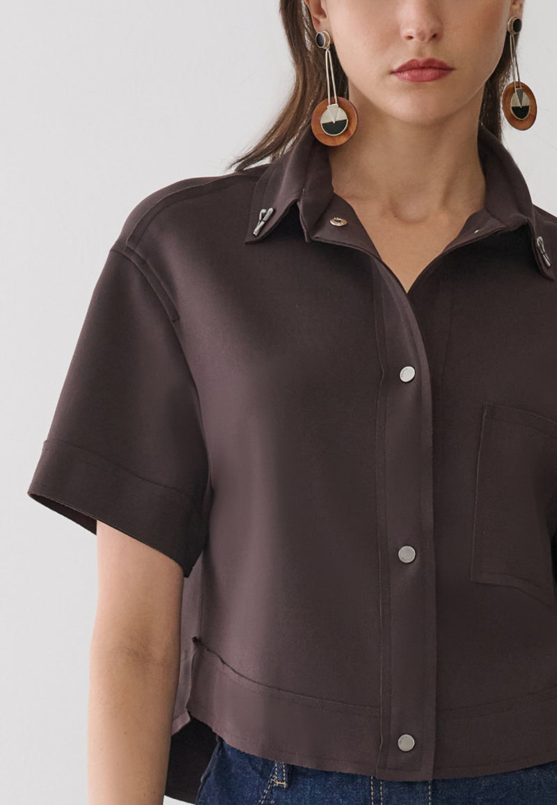 Modelo veste uma camisa marrom de mangas curtas com abotoamento frontal por botões de pressão prateados, detalhe de bolso lateral.