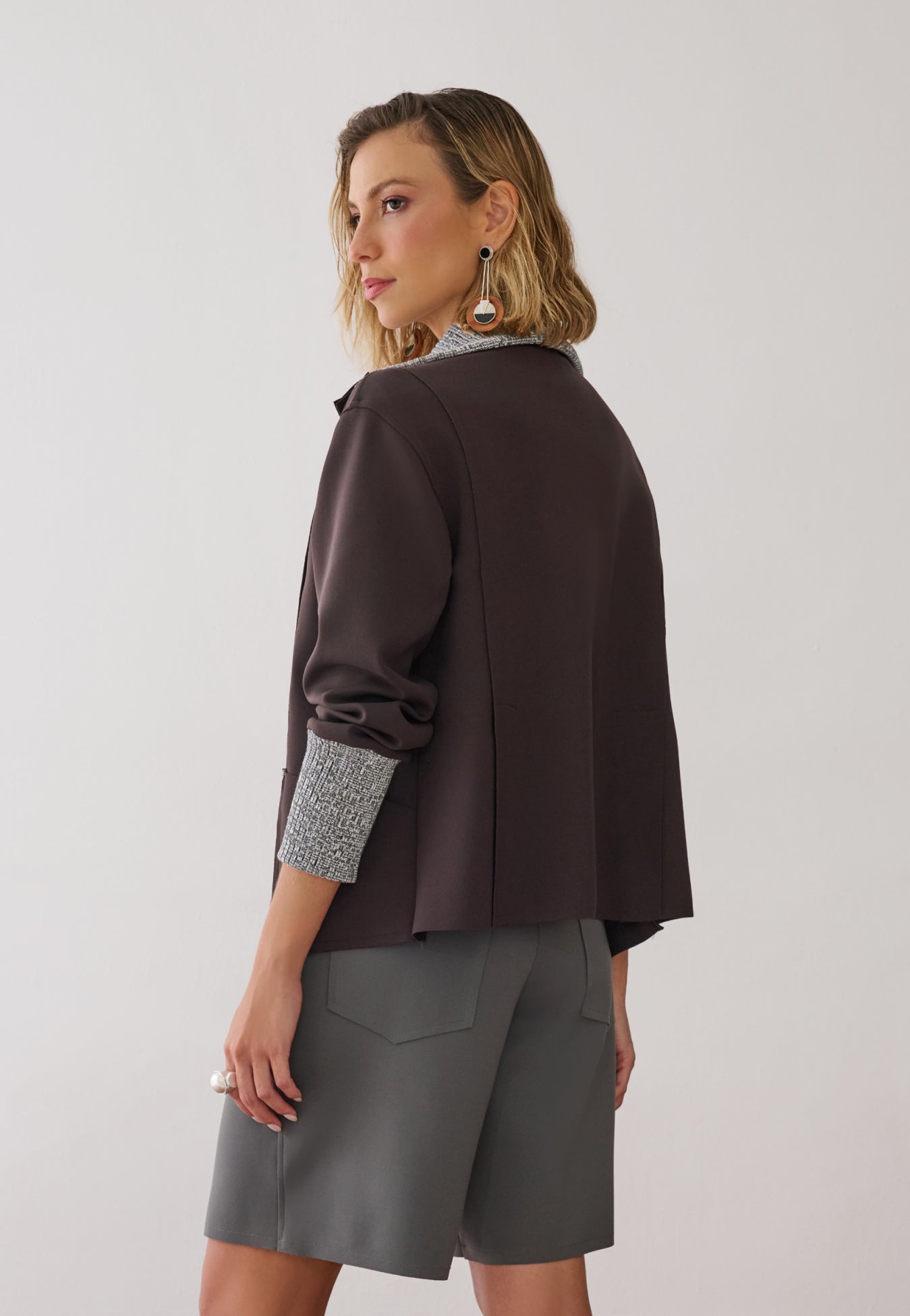 Modelo veste uma jaqueta marrom com zíper frontal, gola e punhos em textura mesclada sobre blusa e bermuda de alfaiataria cinza.