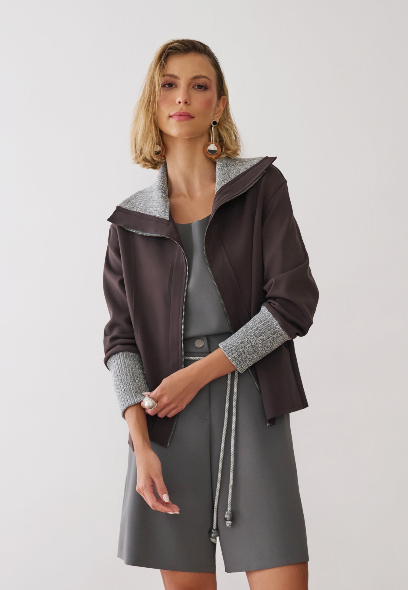 Modelo veste uma jaqueta marrom com zíper frontal, gola e punhos em textura mesclada sobre blusa e bermuda de alfaiataria cinza.