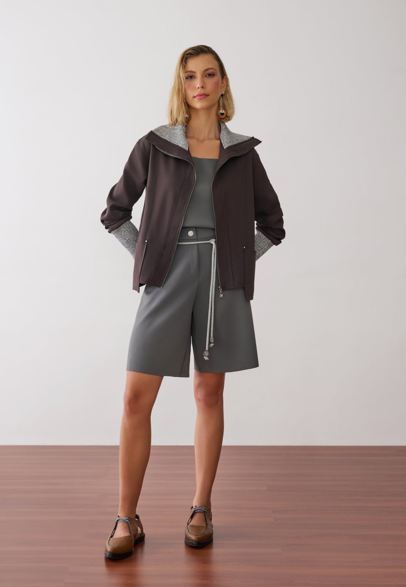 Modelo veste uma jaqueta marrom com zíper frontal, gola e punhos em textura mesclada sobre blusa e bermuda de alfaiataria cinza.