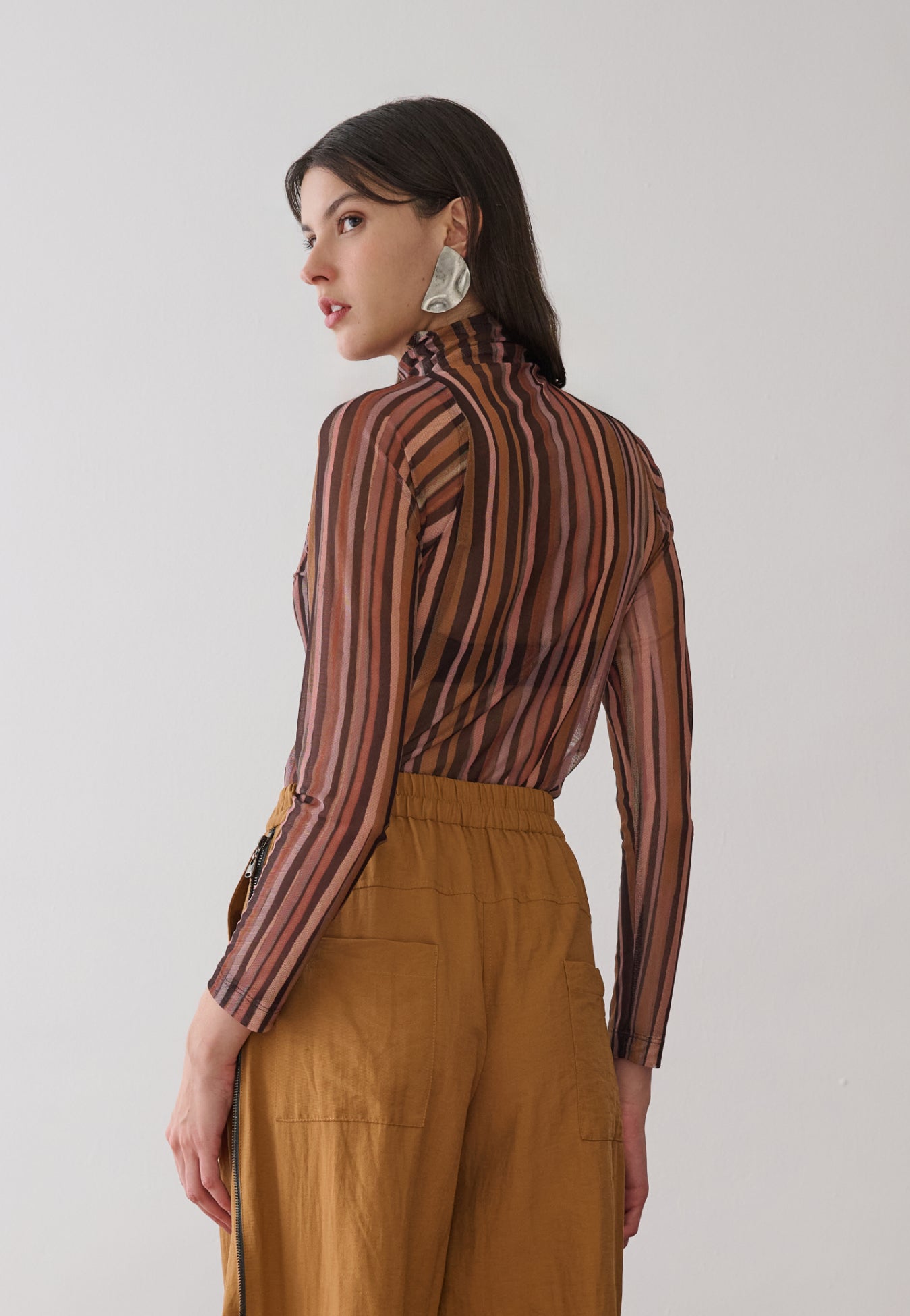 Modelo de costas veste uma blusa de gola alta e mangas longas em tule com estampa de listras verticais multicoloridas, combinada com calças pantalona mostarda de cós elástico e bolsos amplos.