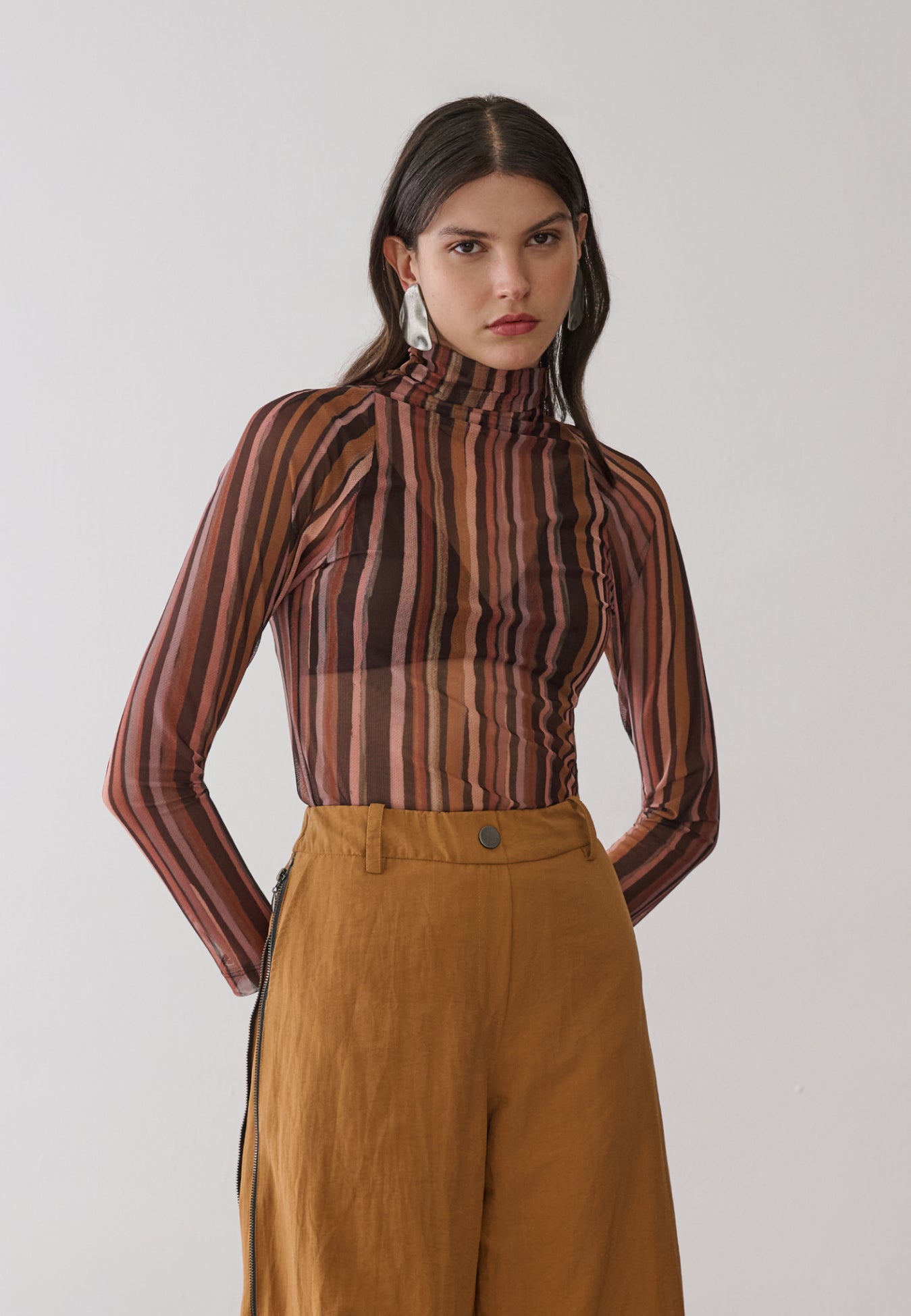 Modelo veste uma blusa de gola alta e mangas longas em tule com estampa de listras verticais multicoloridas, combinada com calças pantalona mostarda.