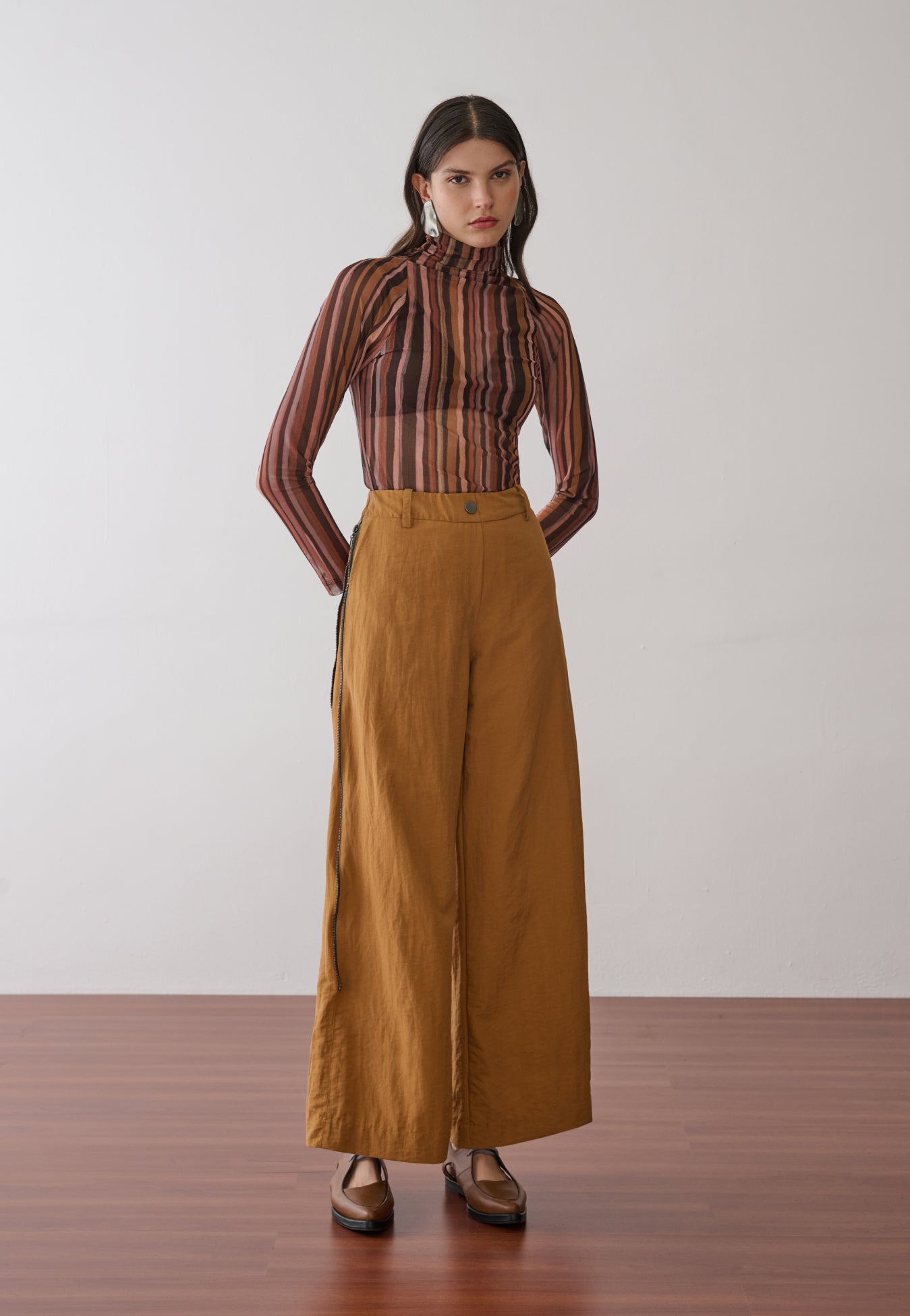 Modelo veste uma blusa de gola alta e mangas longas em tule com estampa de listras verticais multicoloridas, combinada com calças pantalona mostarda e sapatos marrons de bico fino.