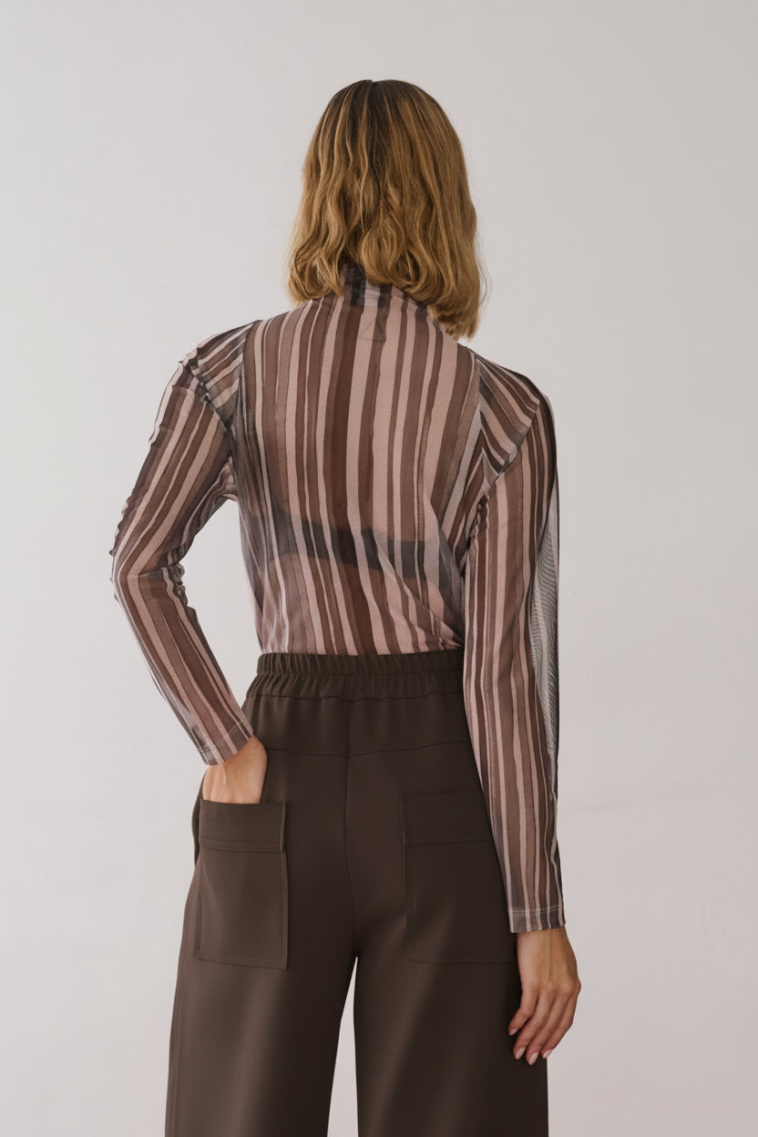 Modelo de costas veste uma blusa de gola alta e mangas longas em tule com estampa de listras verticais em tons de off e cinza, combinada com calças de alfaiataria marrom de cós elástico e bolsos amplos.